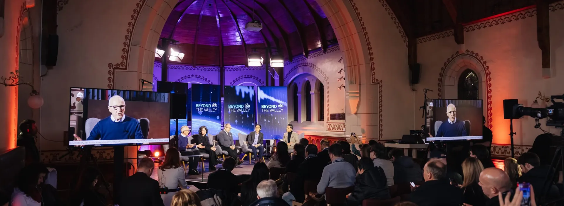 An audience watching a panel discussion inside a vaulted conference hall.