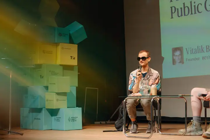 A speaker wearing sunglasses addresses an audience from a conference stage.