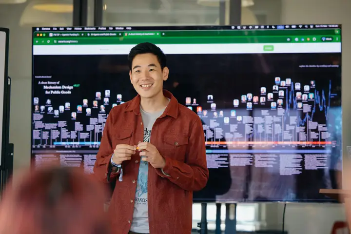 A speaker presents in front of a screen filled with a network diagram.