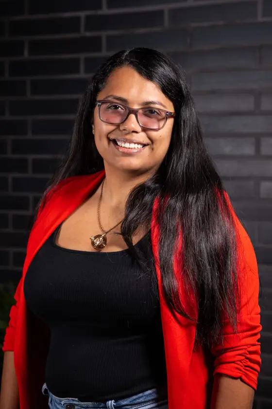 Portrait of a woman wearing glasses and a red jacket against a brick wall.