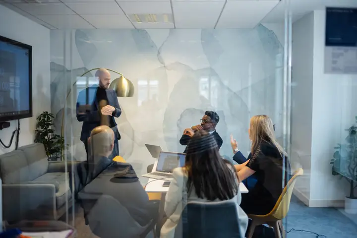 A team listens to a presentation in a glass-walled meeting room.