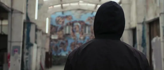 Une silhouette encapuchonnée filmée de dos et de près, occupant environ la moitié droite du cadre — la fresque visible à mi-distance, au centre gauche, au fond de la fonderie